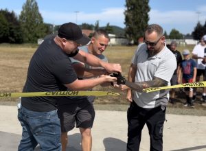 Three men hold giant scissors to cut yellow caution tape.